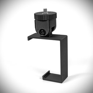 May include: Black metal camera mount with a threaded screw on top. The mount is shaped like a C and has a flat base.