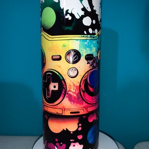 May include: A colorful, abstract design of a video game controller on a black and white background. The controller is painted in shades of yellow, orange, red, green, and blue. The background is covered in black and white paint splatters.