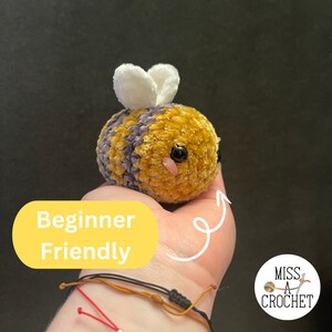 Pode incluir: Um brinquedo de amigurumi de abelha crochetado com listras amarelas e roxas, asas brancas e olhos pretos. A abelha est&aacute; sendo segurada em uma m&atilde;o com o texto "Beginner Friendly" em um fundo amarelo.