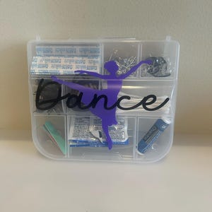 May include: A clear plastic organizer with multiple compartments, containing bandages, cotton swabs, and other small items. A purple silhouette of a ballerina and the word "Dance" are on the front. The organizer is on a white surface.