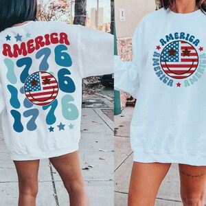 Pode incluir: Camisola branca com um design de cara sorridente vermelho, branco e azul com estrelas e o texto "AMERICA 1776" nas costas. A frente da camisola tem um design semelhante com o texto "AMERICA" repetido em torno da cara sorridente.