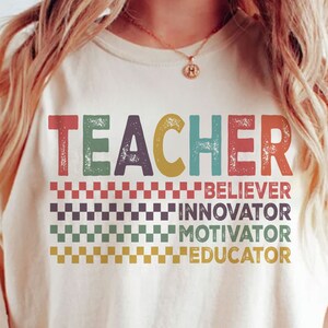 Peut inclure: Un t-shirt blanc avec le mot "TEACHER" en lettres r&eacute;tro color&eacute;es. Sous le mot se trouvent les mots "BELIEVER", "INNOVATOR", "MOTIVATOR" et "EDUCATOR" dans un motif &agrave; carreaux.