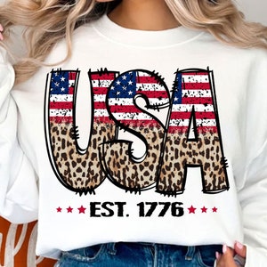 May include: White sweatshirt with a graphic of the word "USA" in red, white, and blue with a leopard print overlay. The graphic also includes the text "EST. 1776" in red.