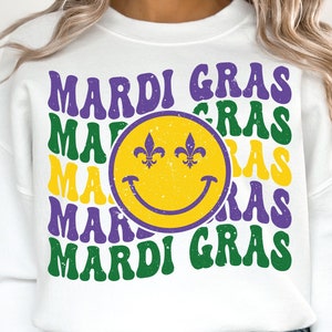 May include: White sweatshirt with a purple and green "Mardi Gras" design featuring a yellow smiley face with fleur-de-lis eyes.