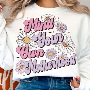 May include: White sweatshirt with a pink and purple graphic design that says "Mind Your Own Motherhood" surrounded by white daisies.