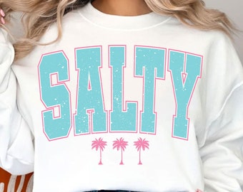 Op de afbeelding: Witte trui met een blauwe en roze grafische afbeelding met de tekst "SALTY" met daaronder drie roze palmbomen.