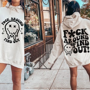May include: A cream colored hoodie with a black graphic that says "F*ck Around Find Out!" with a dripping smiley face. The hoodie has a hood and a kangaroo pocket.