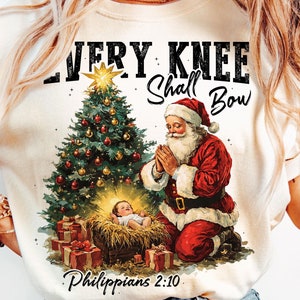 May include: Cream-colored t-shirt featuring a Christmas scene with Santa Claus kneeling in prayer beside a decorated Christmas tree and baby Jesus in a manger. The text "EVERY KNEE SHALL BOW" and "Philippians 2:10" are printed on the shirt.