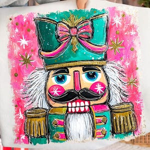 May include: White tote bag featuring a colorful illustration of a nutcracker soldier. The design includes a green hat with a pink bow, a black mustache, and gold accents. Pink and gold stars and a pink background complete the festive design.
