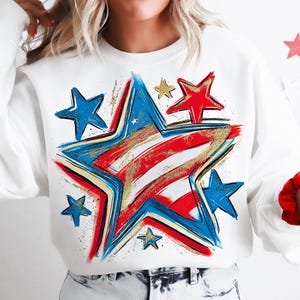 May include: White sweatshirt featuring a large star design in red, white, blue, and gold. The star design has a distressed, painted look, with smaller blue and gold stars scattered around it. A festive, patriotic theme.