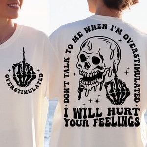 May include: Cream-colored t-shirt with a black graphic of a skeleton hand giving the middle finger, encircled by the text "OVERSTIMULATED." The back features a skull graphic with the text "DON'T TALK TO ME WHEN I'M OVERSTIMULATED. I WILL HURT YOUR FEELINGS."