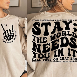 Puede incluir: Sudadera beige con texto gr&aacute;fico e ilustraciones en negro. La parte delantera presenta una mano de esqueleto en un gesto de rock and roll con el texto "THE WORLDS NEEDS YOU". La parte trasera muestra las palabras "STAY; THE WORLD NEEDS YOU IN IT" con un n&uacute;mero de tel&eacute;fono.