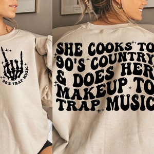 May include: Beige sweatshirt with black text and graphics. The front features a skeleton hand with the words "90's Trap Music." The back says "She cooks to 90's country & does her makeup to trap music."