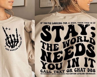 Puede incluir: Sudadera beige con texto gráfico e ilustraciones en negro. La parte delantera presenta una mano de esqueleto en un gesto de rock and roll con el texto "THE WORLDS NEEDS YOU". La parte trasera muestra las palabras "STAY; THE WORLD NEEDS YOU IN IT" con un número de teléfono.