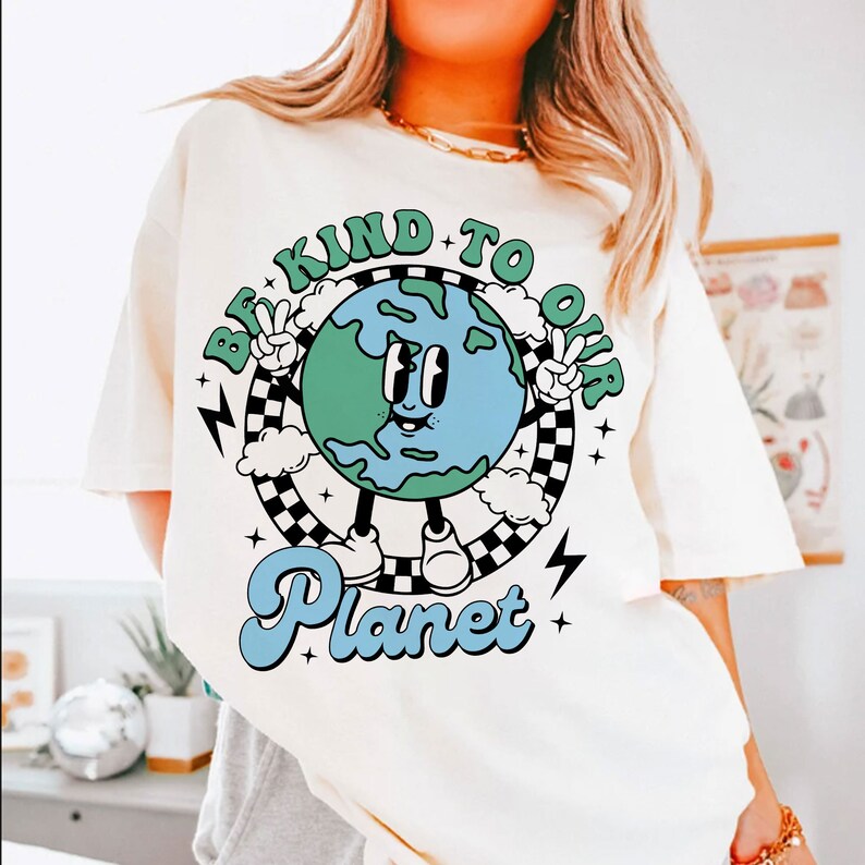 May include: White t-shirt with a graphic of a cartoon Earth with a smiling face and the text "Be Kind To Our Planet" in a retro style.