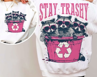 May include: White sweatshirt with a pink recycling bin and three raccoons wearing sunglasses. The text "STAY TRASHY" is printed in pink above the raccoons.