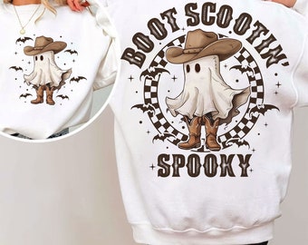 Peut inclure: Sweat-shirt blanc avec un fantôme de dessin animé portant un chapeau de cowboy et des bottes. Le motif comprend des chauves-souris et le texte "Boot Scootin' Spooky". Une version plus petite du motif est sur le devant.