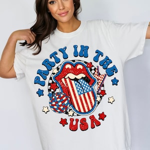 May include: White t-shirt with a graphic of a red, white, and blue tongue with stars and the text "Party in the USA".