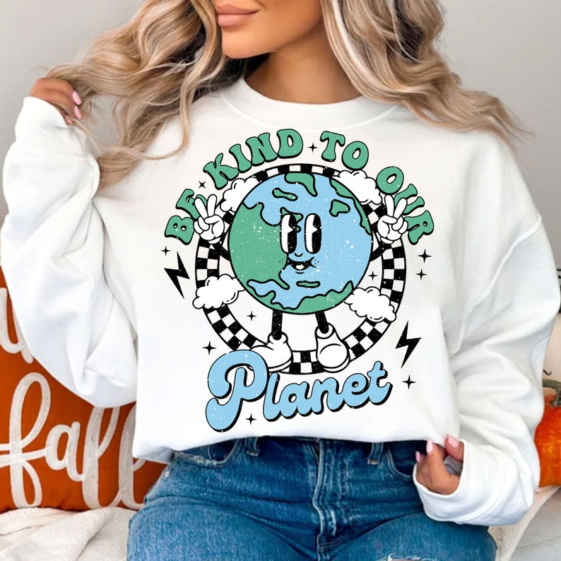 May include: White sweatshirt with a graphic of a cartoon Earth holding up peace signs. The Earth is blue and green with a smiling face. The text "Be Kind To Our Planet" is written in a retro font.
