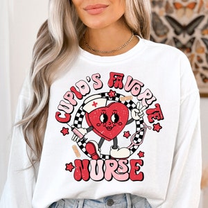 Op de afbeelding: Witte longsleeve trui met een rode hart graphic. Het hart heeft een gezicht en houdt een vredesteken vast. De tekst "Cupid's Favorite Nurse" is in roze en wit rond het hart geschreven.