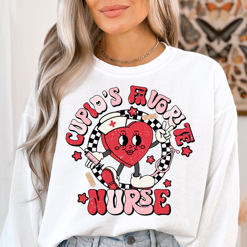 Op de afbeelding: Witte longsleeve trui met een rode hart graphic. Het hart heeft een gezicht en houdt een vredesteken vast. De tekst "Cupid's Favorite Nurse" is in roze en wit rond het hart geschreven.