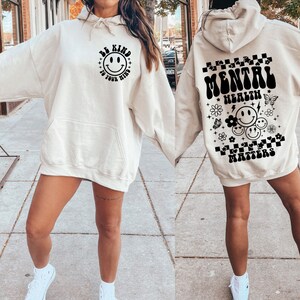 May include: A cream-colored hoodie with a black graphic on the front that says "Be Kind to Your Mind" with a smiley face. The back of the hoodie has a black graphic that says "Mental Health Matters" with smiley faces and flowers.