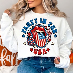 May include: White sweatshirt with a red, white, and blue graphic of a tongue sticking out with a disco ball and stars. The text on the graphic says "Party in the USA".
