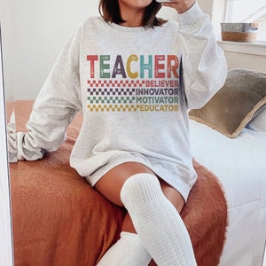Peut inclure: Un sweat-shirt gris avec le mot "TEACHER" en lettres color&eacute;es et un motif &agrave; carreaux. Les mots "BELIEVER", "INNOVATOR", "MOTIVATOR" et "EDUCATOR" sont imprim&eacute;s sous le mot "TEACHER".