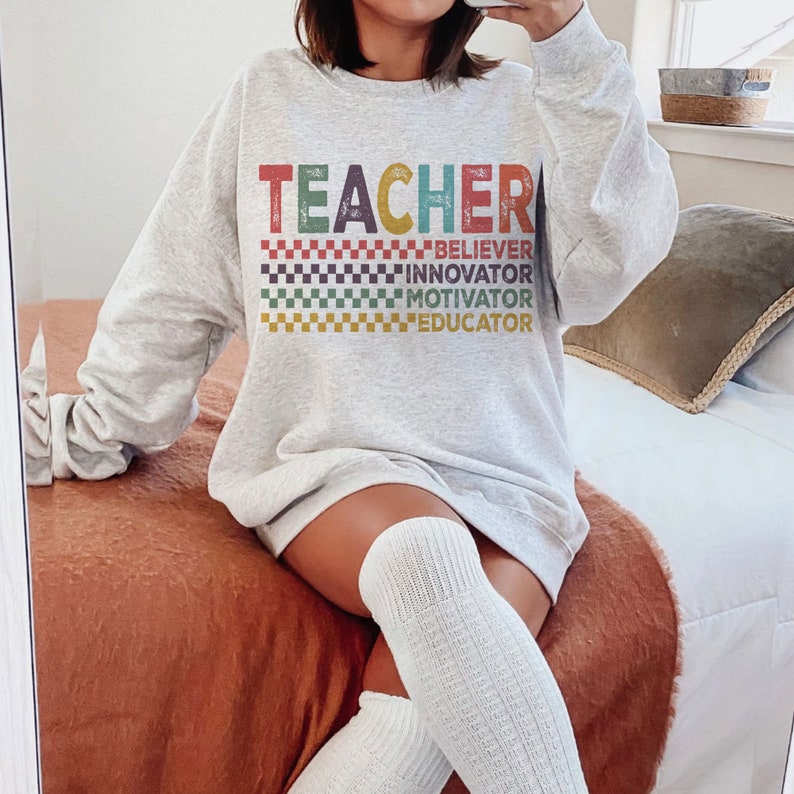 Peut inclure: Un sweat-shirt gris avec le mot "TEACHER" en lettres color&eacute;es et un motif &agrave; carreaux. Les mots "BELIEVER", "INNOVATOR", "MOTIVATOR" et "EDUCATOR" sont imprim&eacute;s sous le mot "TEACHER".