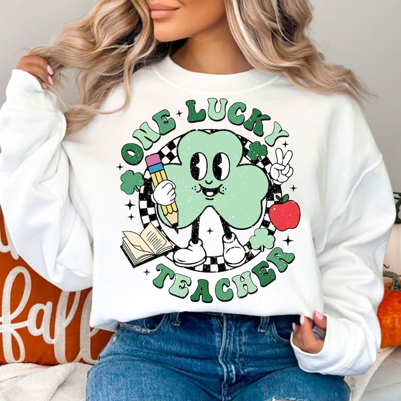 Pu&ograve; includere: Felpa bianca con un trifoglio verde che tiene una matita e una mela. Il trifoglio ha un viso e indossa delle scarpe. Il testo intorno al trifoglio dice "One Lucky Teacher".