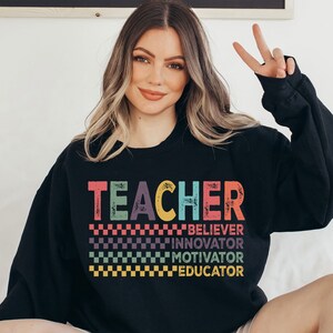 Peut inclure: Sweat-shirt noir avec le texte "TEACHER" en couleurs arc-en-ciel et les mots "BELIEVER", "INNOVATOR", "MOTIVATOR" et "EDUCATOR" en dessous.