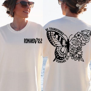 May include: White t-shirt with black text and a butterfly graphic on the back. The text reads "BE TRANSFORMED" and "ROMANS 12:2". The front of the shirt has the text "ROMANS 12:2" with a heart symbol.