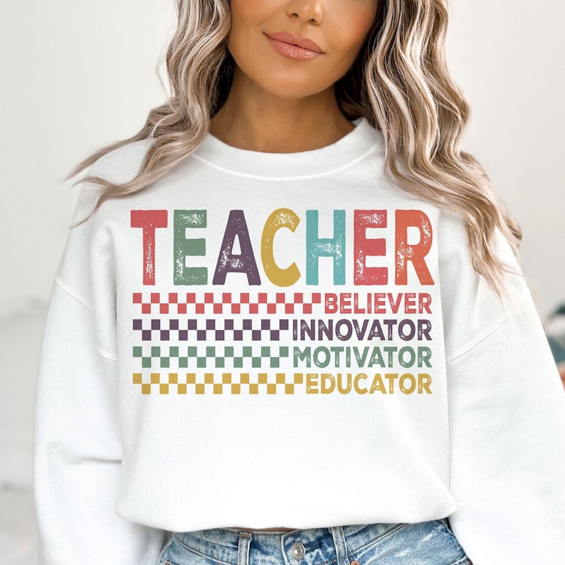Peut inclure: Sweat-shirt blanc avec le mot "TEACHER" en lettres vintage color&eacute;es. En dessous du mot se trouvent les mots "BELIEVER", "INNOVATOR", "MOTIVATOR" et "EDUCATOR" en lettres plus petites.