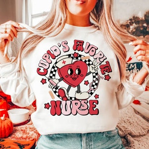 Op de afbeelding: Wit sweatshirt met een rode hartgrafiek waarop in roze en rode letters "Cupid's Favorite Nurse" staat. Het hart draagt een verpleegstersmuts en houdt een spuit.