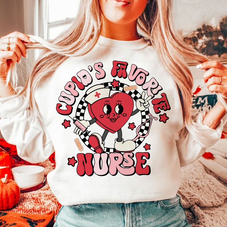Op de afbeelding: Wit sweatshirt met een rode hartgrafiek waarop in roze en rode letters "Cupid's Favorite Nurse" staat. Het hart draagt een verpleegstersmuts en houdt een spuit.