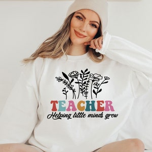 以下が含まれることがあります： 虹色の文字で「TEACHER Helping little minds grow」と書かれた、黒い花柄の白いスウェットシャツです。