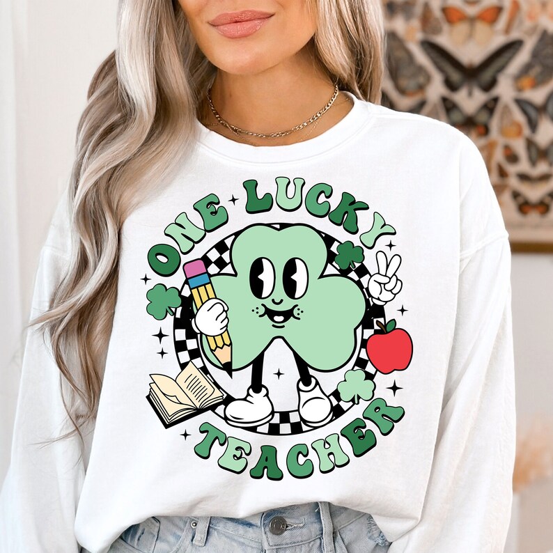 Pu&ograve; includere: Felpa bianca a maniche lunghe con un personaggio di cartone animato a forma di trifoglio verde che tiene una matita e una mela. Il testo "One Lucky Teacher" &egrave; scritto intorno al personaggio.