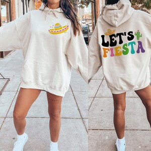 May include: A cream-colored hoodie with a colorful "Let's Fiesta" graphic on the back and a sombrero graphic on the front. The hoodie has a drawstring hood and a kangaroo pocket.