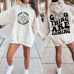 K&ouml;nnte beinhalten: Ein cremefarbener Kapuzenpullover mit dem Text "Good Things Are Coming" in Schwarz, mit einem Smiley-Blumen-Motiv auf der Vorderseite und zwei Smileys auf der R&uuml;ckseite.