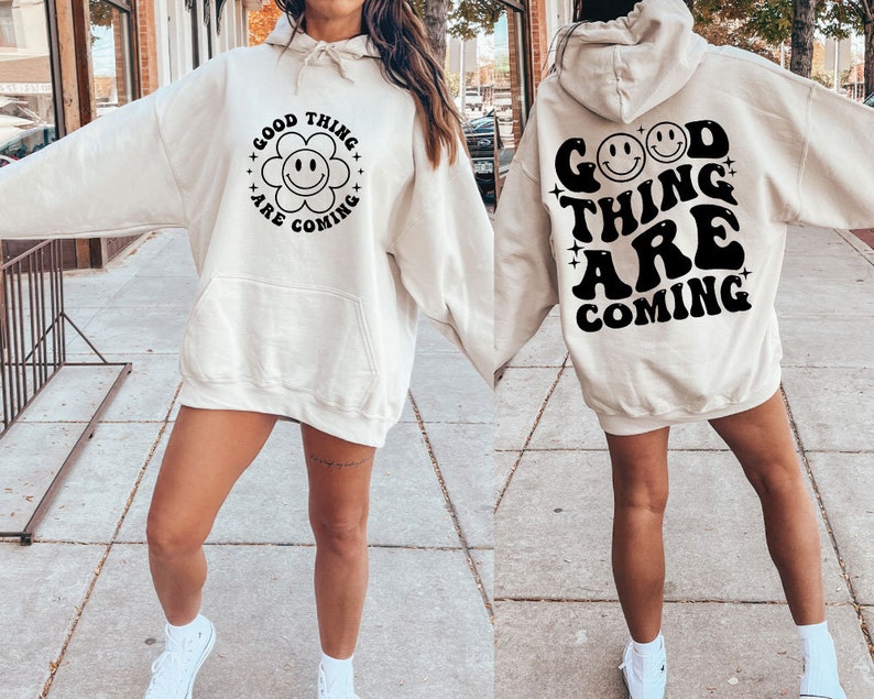 K&ouml;nnte beinhalten: Ein cremefarbener Kapuzenpullover mit dem Text "Good Things Are Coming" in Schwarz, mit einem Smiley-Blumen-Motiv auf der Vorderseite und zwei Smileys auf der R&uuml;ckseite.