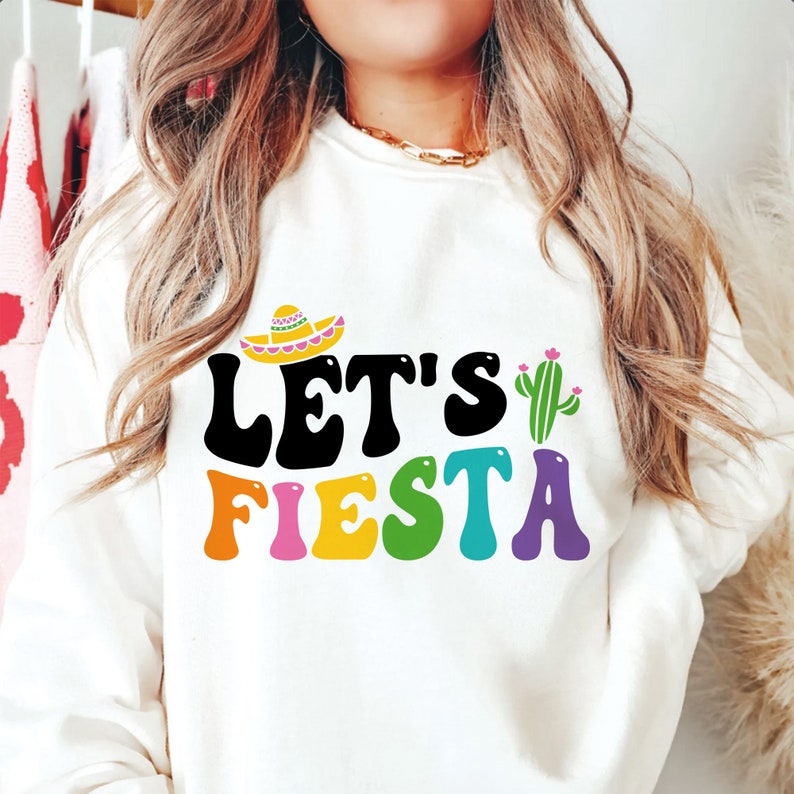 May include: White sweatshirt with the text "Let's Fiesta" in colorful letters. A sombrero and cactus are above the text.