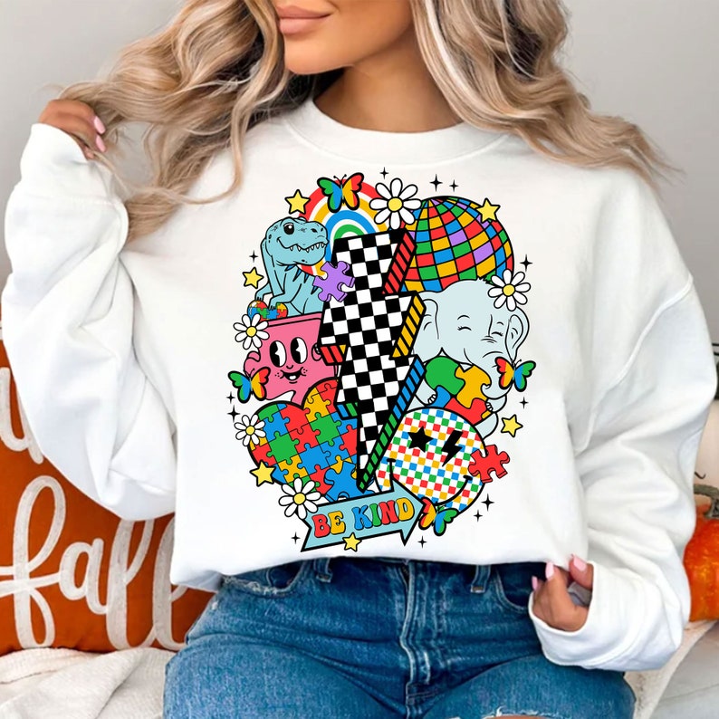 Op de afbeelding: Witte trui met een kleurrijke grafische print met een bliksem, een dinosaurus, een olifant, een puzzelhart, een regenboog, een discobal en vlinders. De tekst "Be Kind" is onder de print gedrukt.