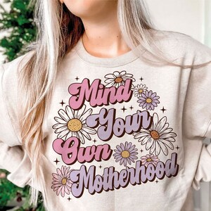 May include: A beige sweatshirt with the text "Mind Your Own Motherhood" in pink and purple lettering with a daisy flower design.