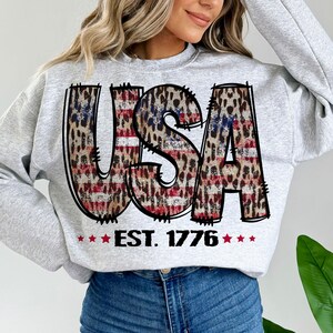 May include: A gray sweatshirt with a graphic of the word "USA" in red, white, and blue with a leopard print pattern. The text "EST. 1776" is below the graphic.