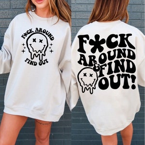 May include: A white sweatshirt with a black graphic print on the front and back. The front graphic says "F*CK AROUND FIND OUT" with a dripping smiley face. The back graphic says "F*CK AROUND & FIND OUT!" with a dripping smiley face.