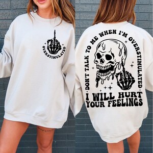 May include: White sweatshirt with a black skull and middle finger graphic on the back. The text reads "Don't talk to me when I'm overstimulated. I will hurt your feelings."