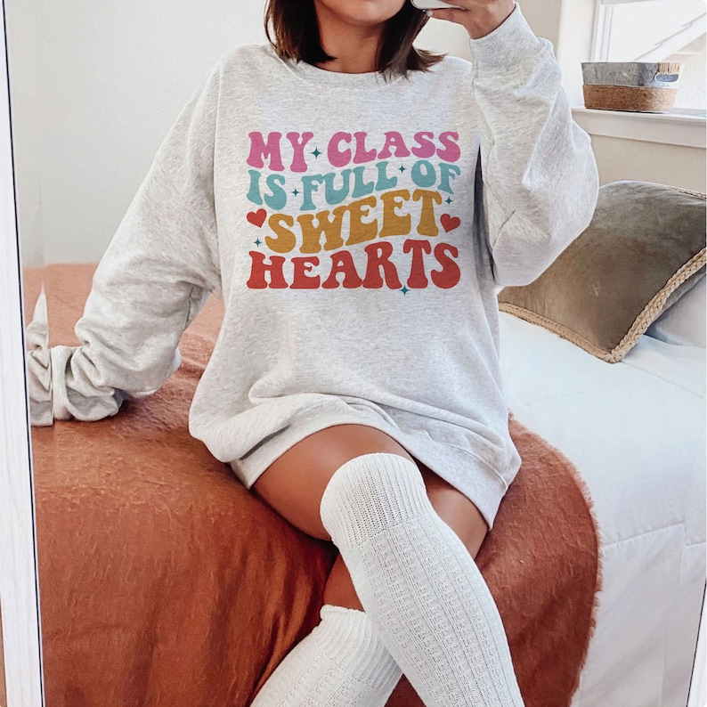 以下が含まれることがあります： ハートと星が散りばめられたカラフルなレトロなフォントで「My Class is Full of Sweet Hearts」と書かれたグレーのスウェットシャツです。