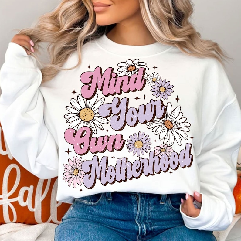 May include: White sweatshirt with a pink and purple graphic design that says "Mind Your Own Motherhood" surrounded by white daisies.