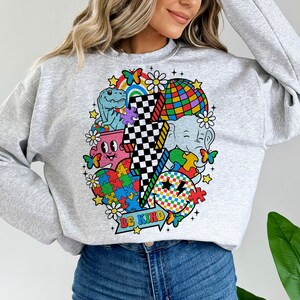 Op de afbeelding: Een grijs sweatshirt met een kleurrijke grafische print met een bliksem, een dinosaurus, een olifant, een puzzelhart, een smiley en een discobal. De grafiek bevat ook de tekst "Be Kind".