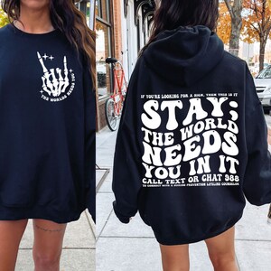 Puede incluir: Sudadera negra con texto blanco. La parte delantera presenta un gesto de mano de esqueleto y las palabras "The Worlds Needs You". La parte trasera de la sudadera con capucha dice "Stay; The World Needs You In It" con detalles de prevenci&oacute;n del suicidio.